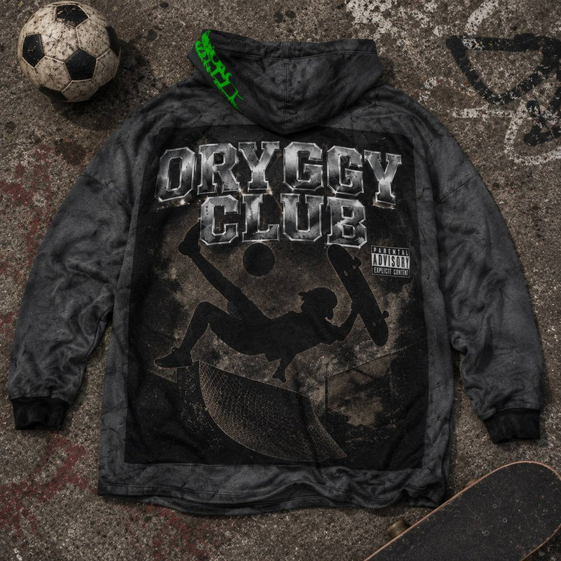 Oryggy Club oversized hooded fleece with skateboarding graphic laid on graffiti-covered concrete, styled with a skateboard and soccer ball in an urban streetwear flat lay.