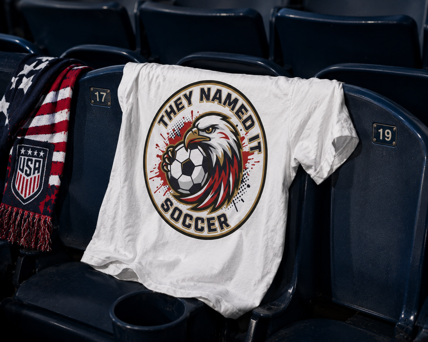 USA soccer fan t-shirt draped over a stadium seat backrest, featuring an eagle and soccer ball graphic, with a USA supporter scarf and soccer ball in an empty stadium section.